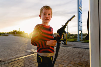 Portrait of boy holding fuel pump at gas station during sunset