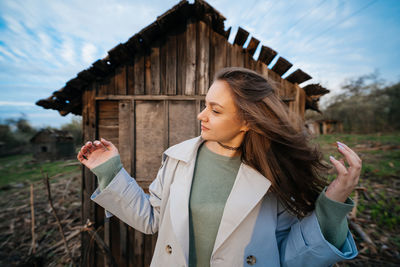 Beautiful girl with long hair in a grey trench coat next to an old wooden house outdoors in spring