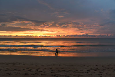 People walking on shore at beach against sky during sunset