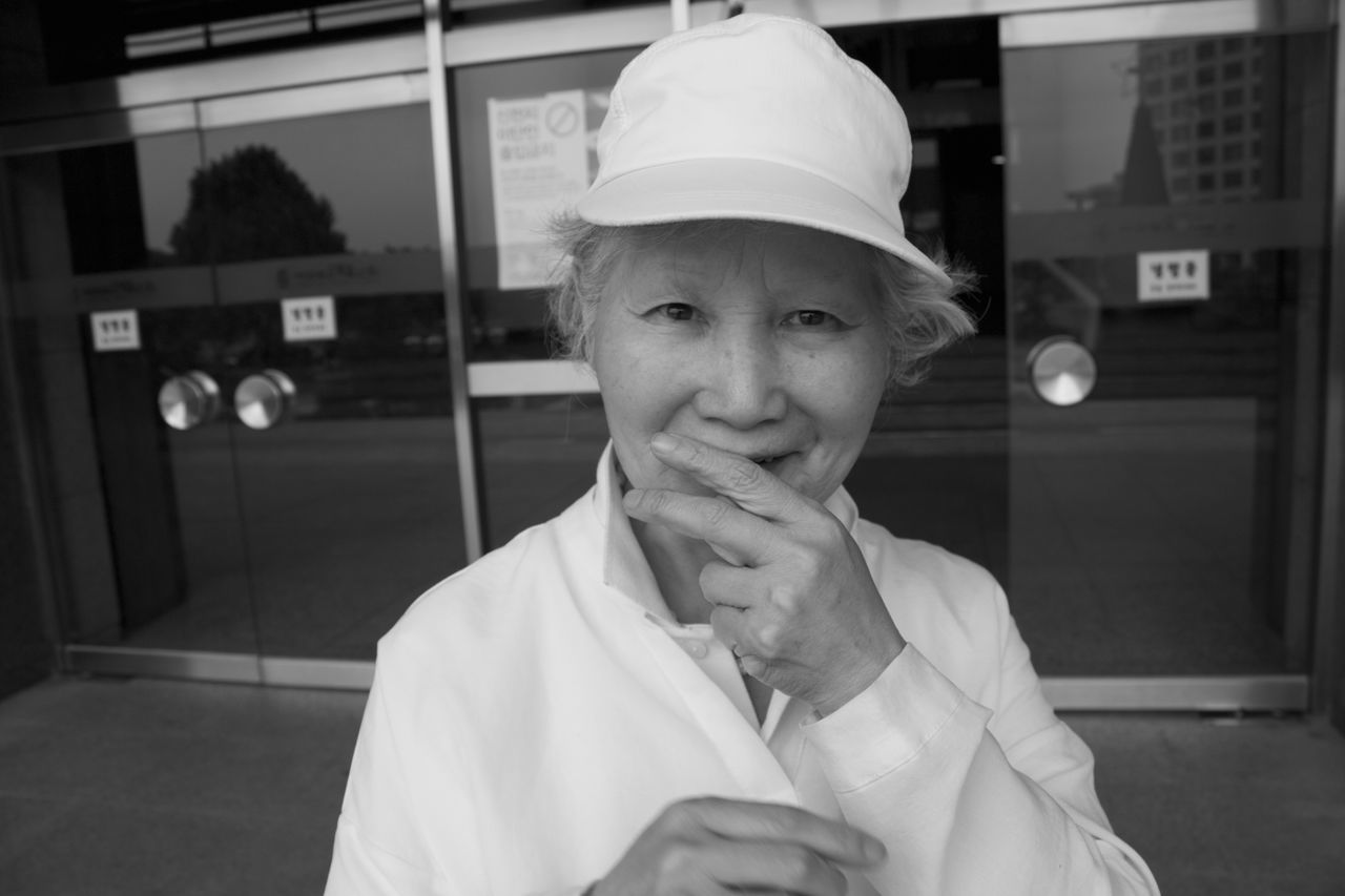 real people, looking at camera, front view, one person, portrait, building exterior, lifestyles, architecture, built structure, young adult, cap, standing, outdoors, headwear, day, young women