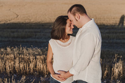 Smiling man touching pregnant girlfriend belly while standing on field