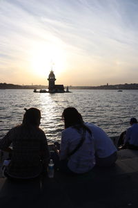 Rear view of people sitting on sea against sky during sunset