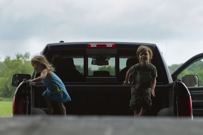 Siblings standing in pick-up truck against cloudy sky