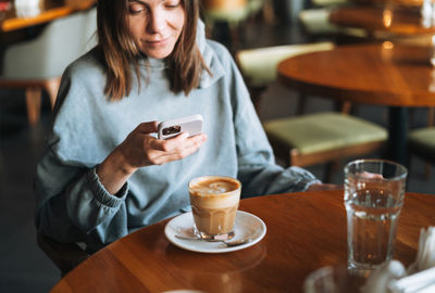 Young brunette woman wearing casual longsleeve with mobile phone in hands with cup of coffee in cafe
