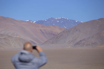Rear view of man photographing on mobile phone against mountains and clear sky