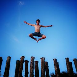 Low angle view of shirtless teenage boy jumping over wooden posts against clear blue sky