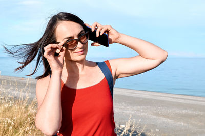 Beautiful woman in sun glasses and red top talking on cell phone on the beach with sea in background