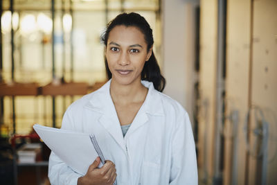 Portrait of confident young chemistry student wearing lab coat standing with book in university