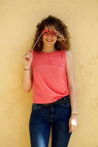 Portrait of smiling young woman holding eye mask prop while standing against wall