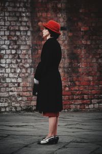 Side view of woman standing on footpath against brick wall