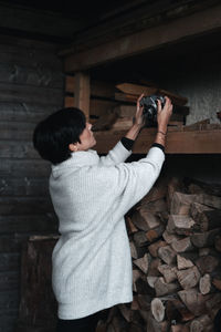 Woman photographing while holding camera on shelf with firewood