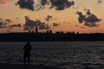 Rear view of silhouette man standing by sea against sky during sunset