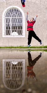 Reflection of boy in pond throwing cap while jumping on field against building