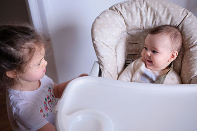 Happy infant is smiling at his sister while sitting in the high chair