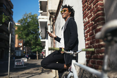 Side view of cheerful man using smart phone while standing by wall