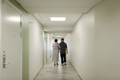 Full length rear view of senior woman walking arm in arm with male nurse at retirement home corridor