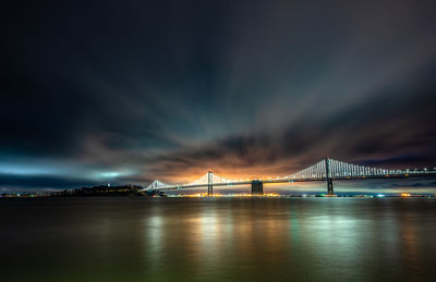 Illuminated bridge over river against cloudy sky