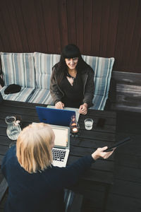 High angle view of woman showing digital tablet to female friend using laptop at table