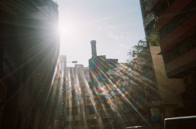 Low angle view of sunlight streaming through buildings against sky