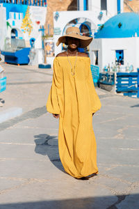 Beautiful african american woman in yellow dress and sun hat walks through streets with summer cafe