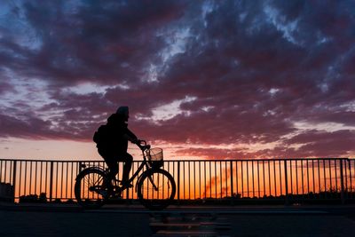 Silhouette man riding bicycle on bridge against sky during sunset