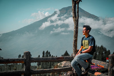 Young man sitting on wood against sky
