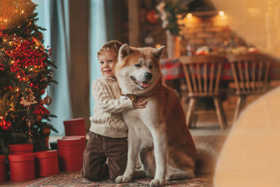 Candid authentic happy little boy in knitted beige sweater hugs dog with bow tie at home on xmas