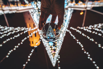 Cropped hand holding crystal ball against illuminated lighting equipment at night