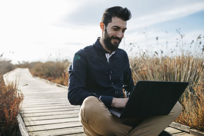 Man using laptop while sitting on boardwalk against sky