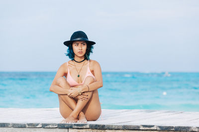 Full length of woman in bikini sitting on pier against sea and sky