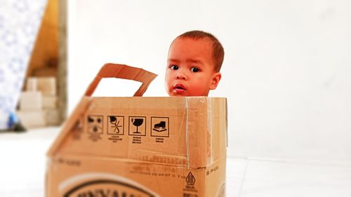 A toddler boy, getting into the box of a toy car, made of cardboard, stares seriously.