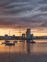 Boats moored in sea by city against cloudy sky during sunset