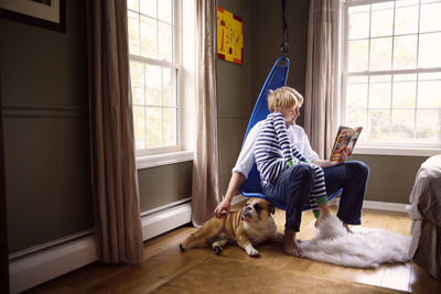 Father reading book to son and stroking dog while sitting on swing at home