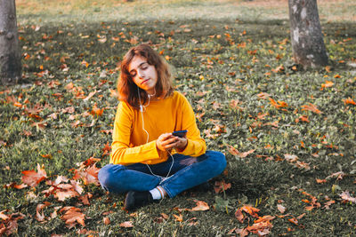 Young girl in yellow sweater listening music with earphones sitting on autumn grass