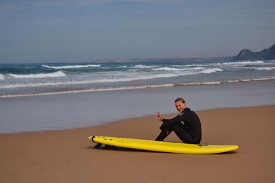 Young woman gesturing peace sign while sitting by surfboard on sea shore