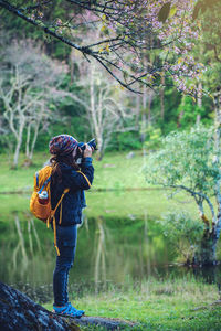 Side view full length of young woman photographing from camera by lake at forest