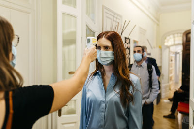 Female with infrared thermometer checking businesswoman's temperature in corridor