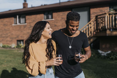 Happy couple holding wineglass while walking at backyard