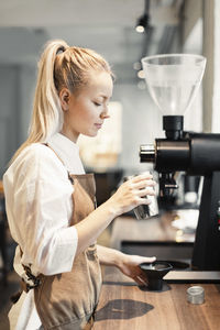 Side view of female barista holding container by coffee maker at counter in cafe