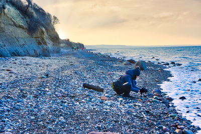 Side view of woman photographing while crouching on pebble stones against sky during sunset