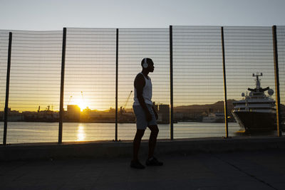 Side view full body of african american athlete standing on seafront while resting after fitness workout at dusk