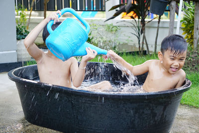 Shirtless boy pouring water on brother while sitting in bucket at yard