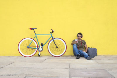Man listening music while sitting by bicycle on footpath against yellow wall