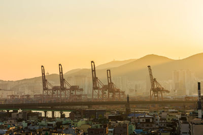 Cranes at harbor against sky during sunset