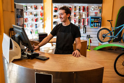 Happy young cashier in black t-shirt smiling and using computers while working in modern bike store