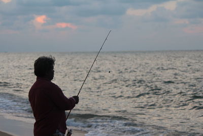 Rear view of man fishing in sea against sky during sunset