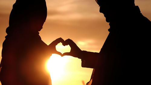 Silhouette couple forming heart shape against orange sky