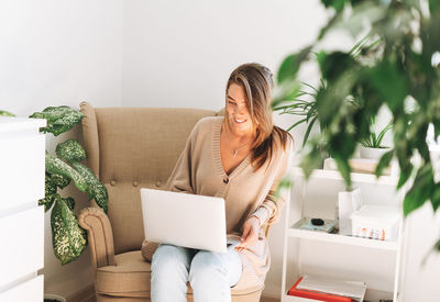 Young smiling attractive woman in cozy beige cardigan working at laptop sitting in chair at home 
