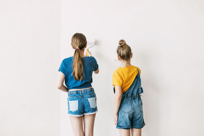 Two sisters are painting the walls of the house white, in denim overalls and a bright yellow t-shirt