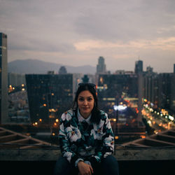 Portrait of young woman sitting against illuminated cityscape at night